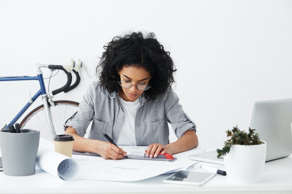 Candid shot of professional skilled Afro American female architect holding ruler and pen while drawi